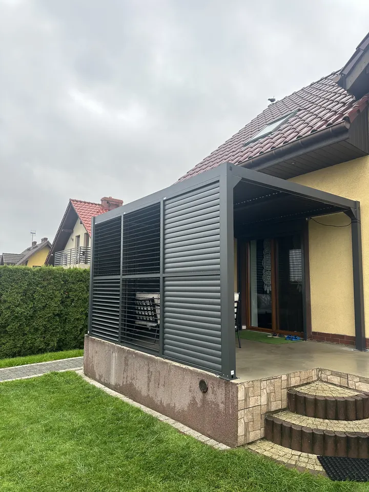 Grafitowa pergola z poziomymi osłonami na betonowym podeście przy żółtym domu w Pawłowicach
