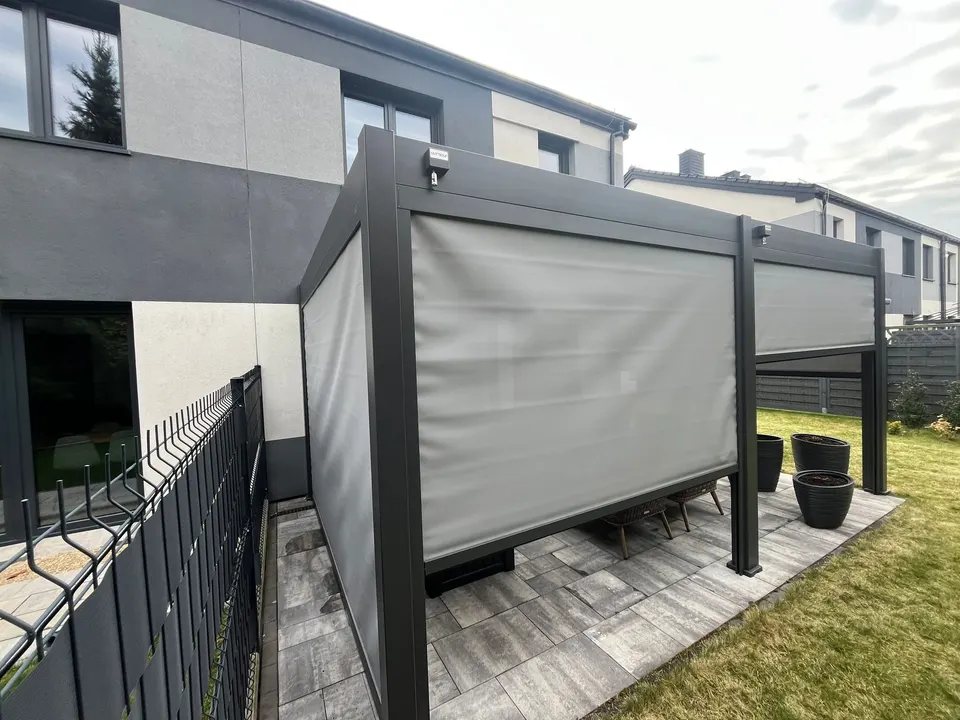 Grafitowa pergola lamelowa w Katowicach z częściowo opuszczonymi jasnymi osłonami i meblami