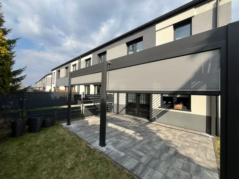 Pergola przy elewacji domu z pionowymi osłonami opuszczonymi do połowy